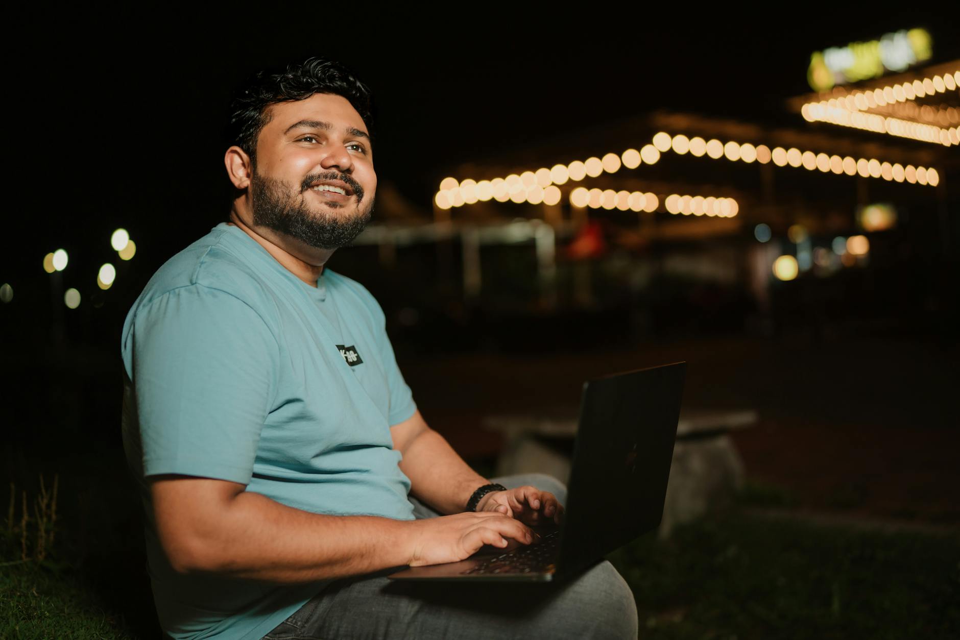 Smiling man typing on laptop outdoors at night with string lights in the background.