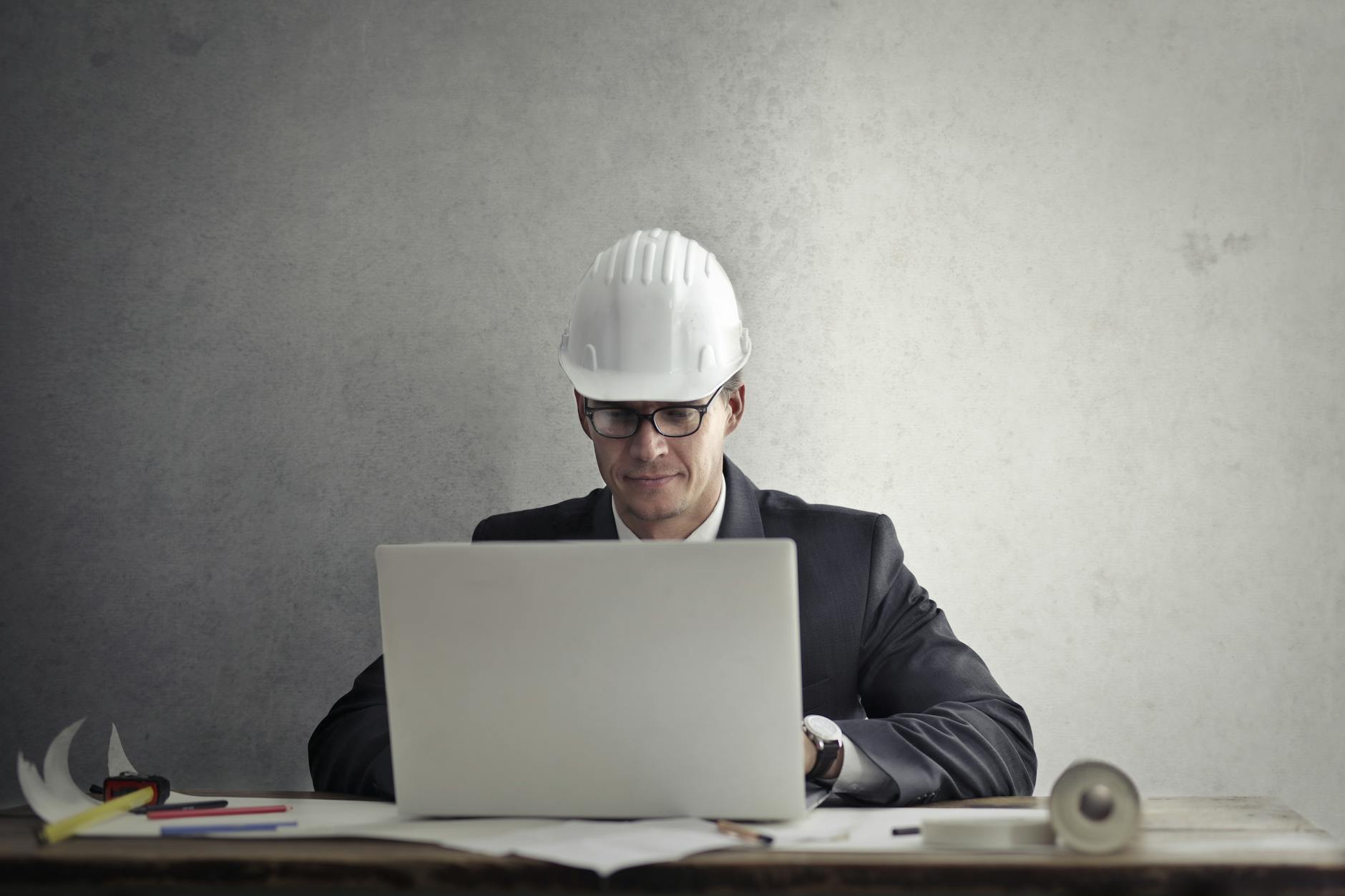 Professional engineer wearing hardhat working on construction plans at desk with laptop in office.