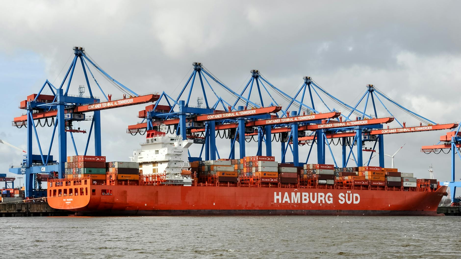 Red container ship docked at Hamburg Terminal under cranes on a cloudy day.