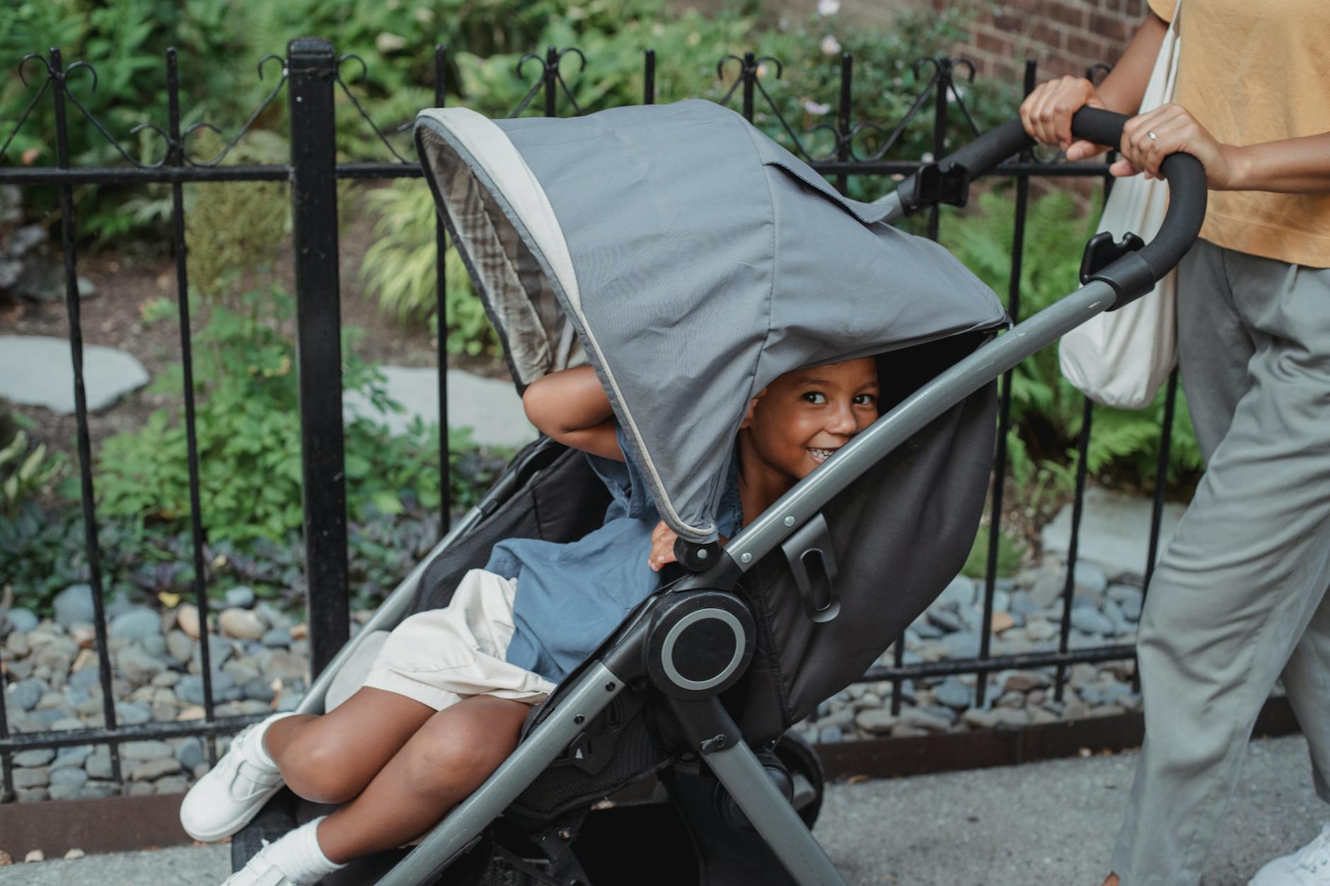 Happy ethnic girl sitting in stroller and smiling while walking on city street with anonymous mother in daytime