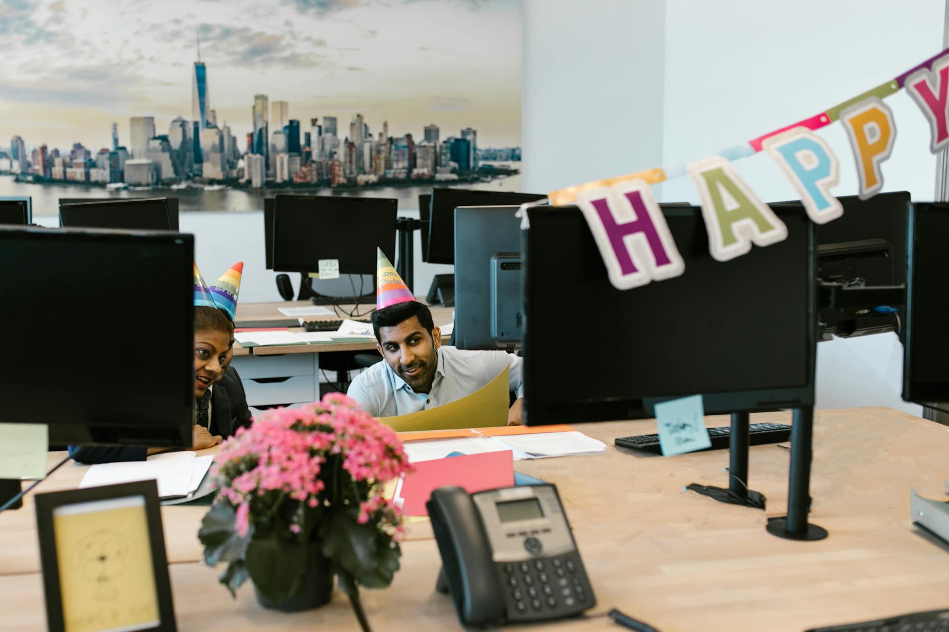 Colleagues wearing party hats celebrate a birthday in a modern office with decorations.