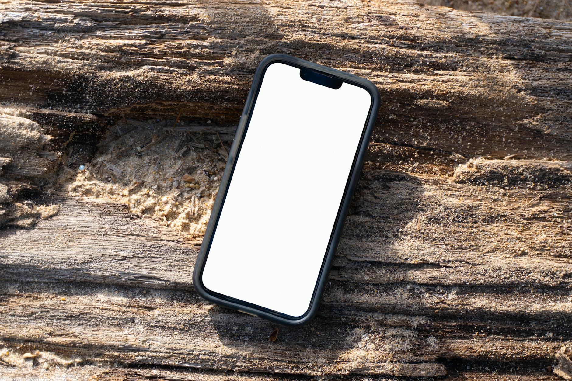 iPhone mockup with blank screen on a sandy wooden log at Kamala Beach, Phuket, Thailand.