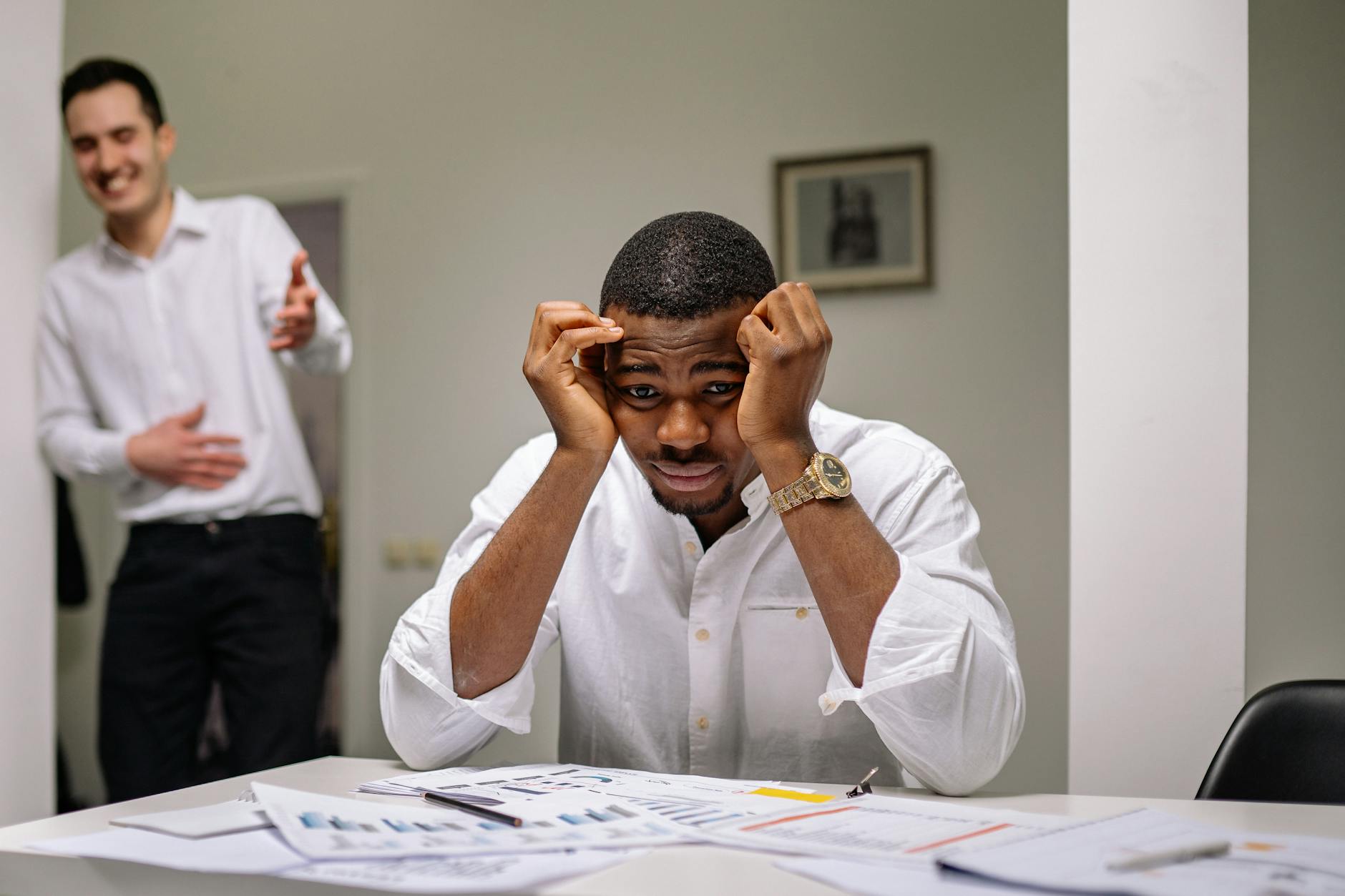 Tensed businessman at desk while coworker is laughing. Office stress concept.
