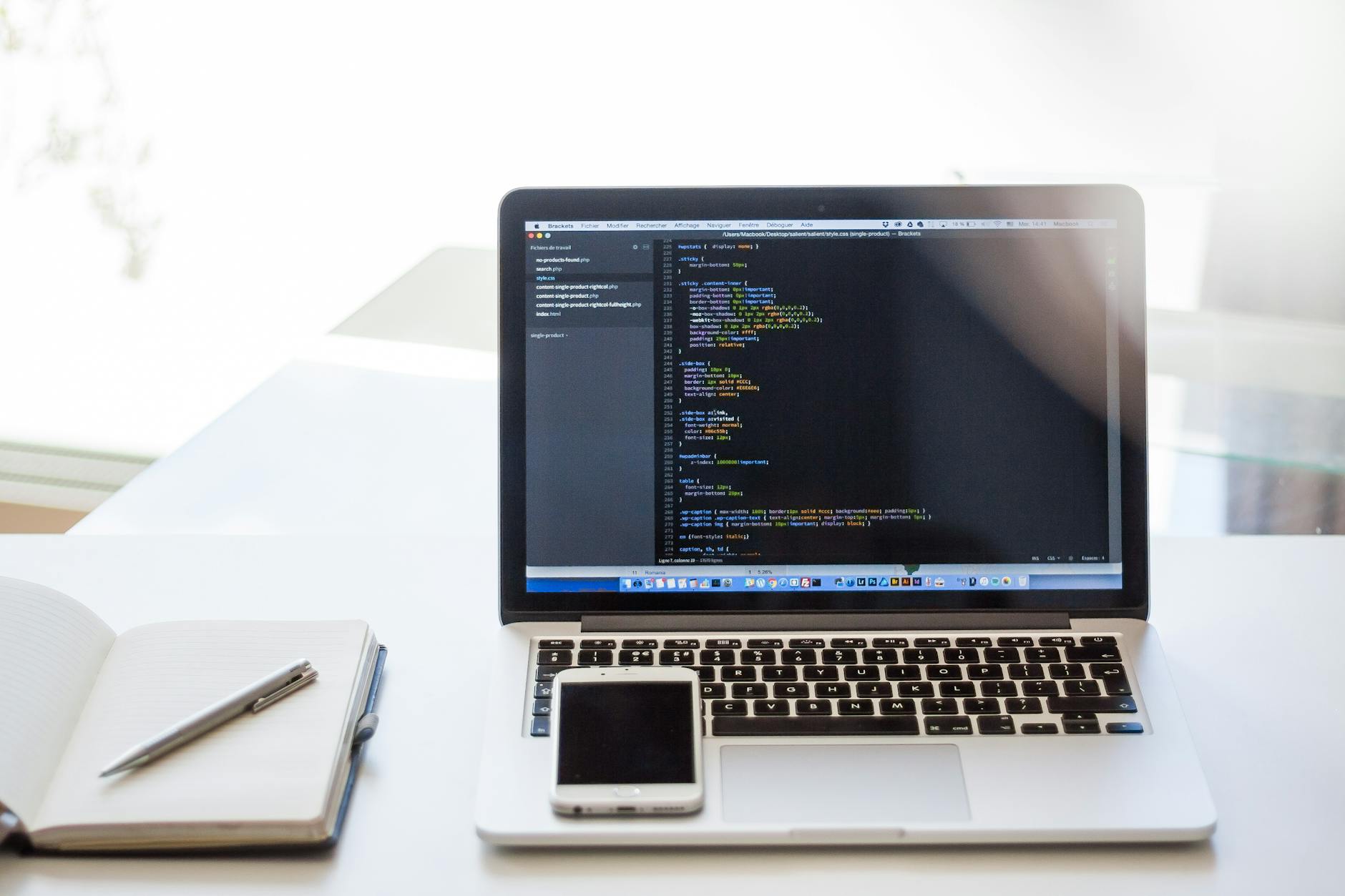 Open laptop displaying code on a desk with smartphone and notebook, ideal for technology and business use.